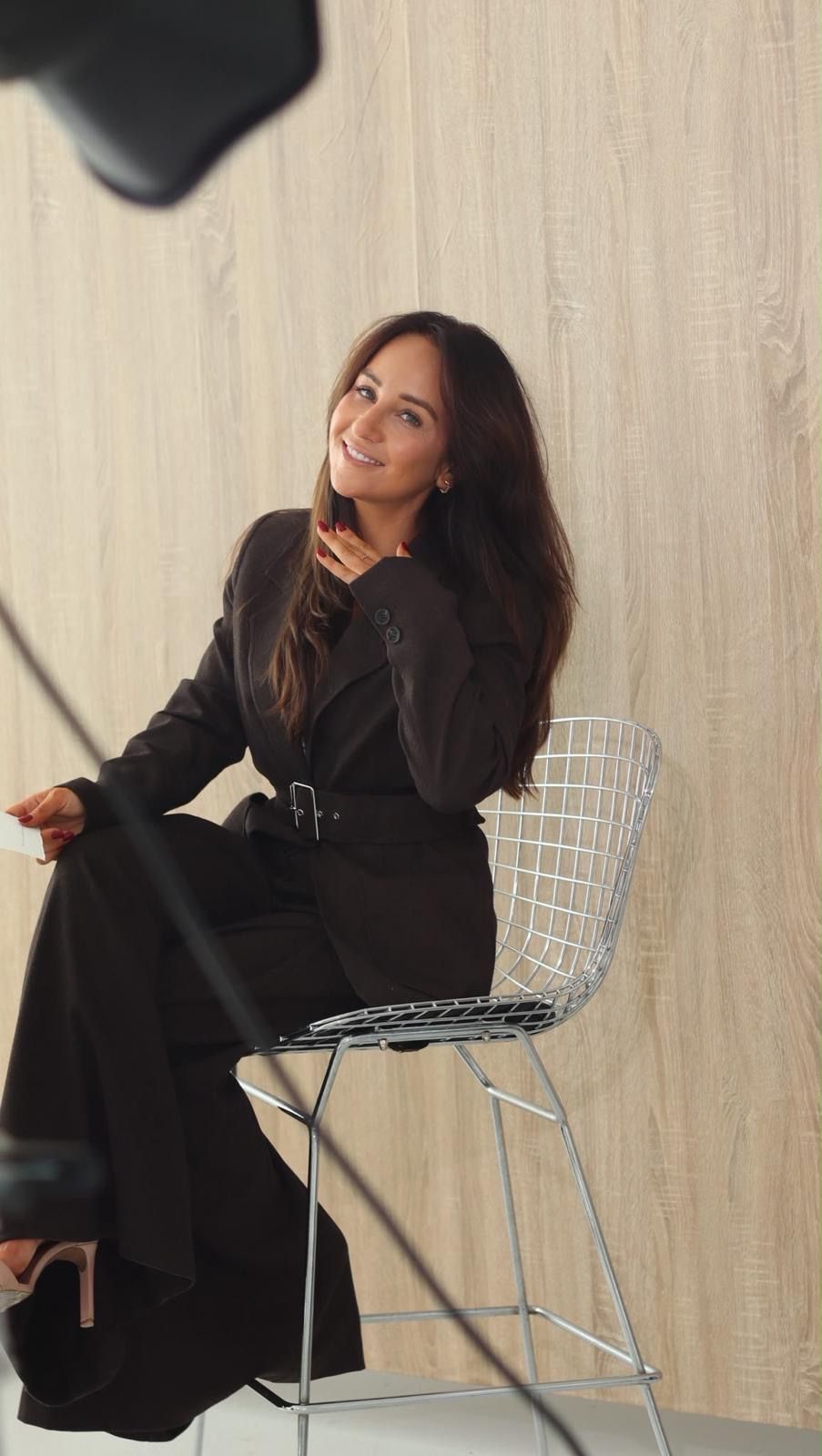 Melissa laughing on set, sitting on a wire chair with a studio light in the foreground