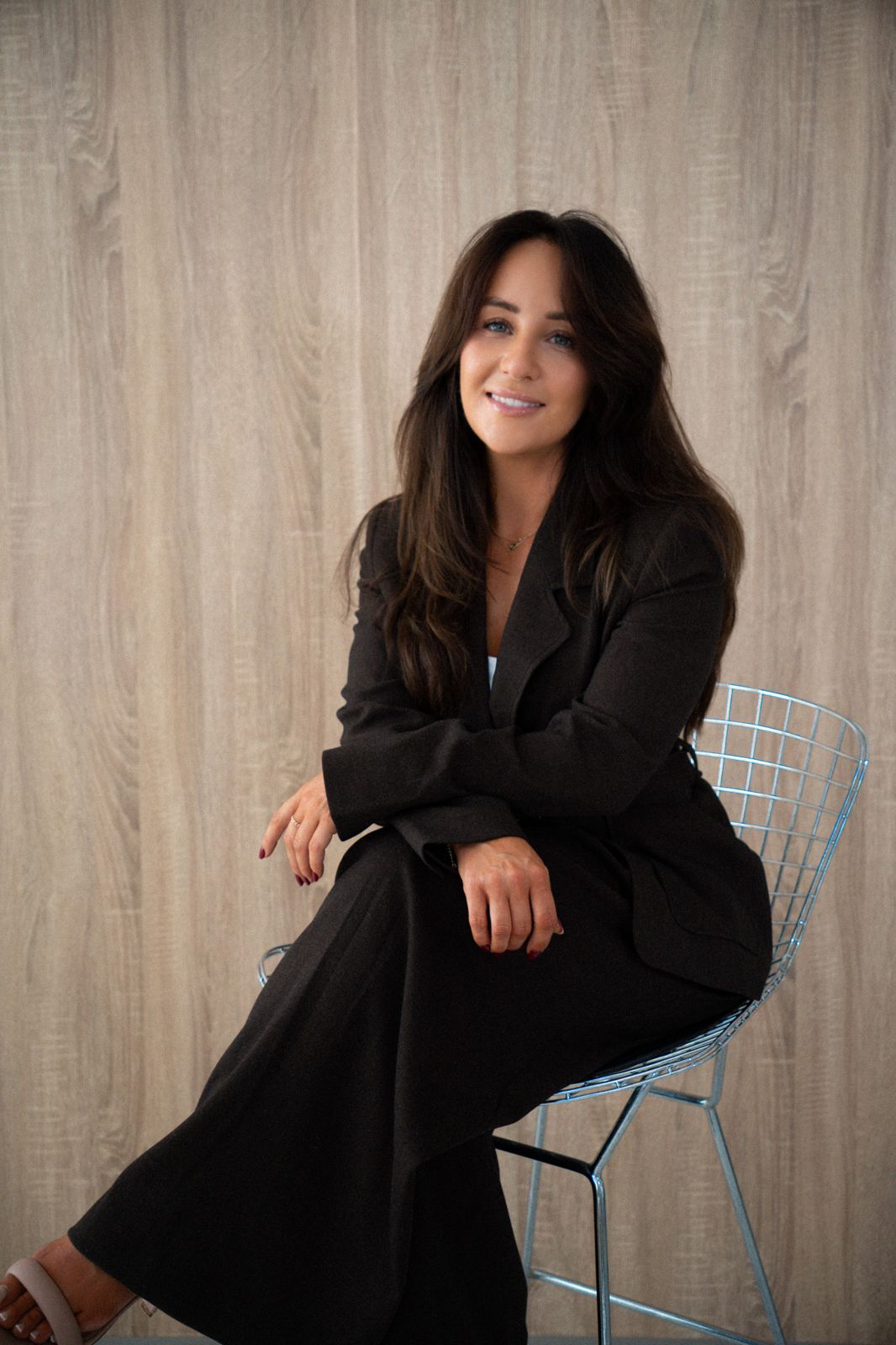 Melissa Mitchelmore, founder of Savoir, sitting on a wire chair in a dark brown suit, smiling